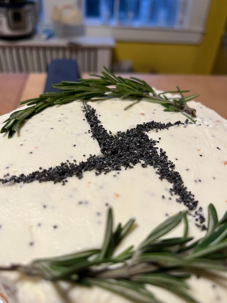 Close up of the top of a frosted cake. There is a Bridhe’s cross picked out in poppyseeds and two stems of rosemary.