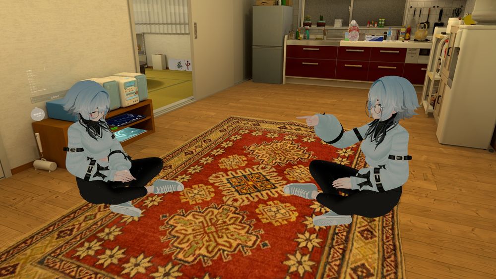 Two identical anime-style characters with short light blue hair, glasses, and matching outfits sit cross-legged on a red patterned rug in a cozy room. One character points at the other, who looks down with hands folded. The background shows a kitchen and a small tatami room.