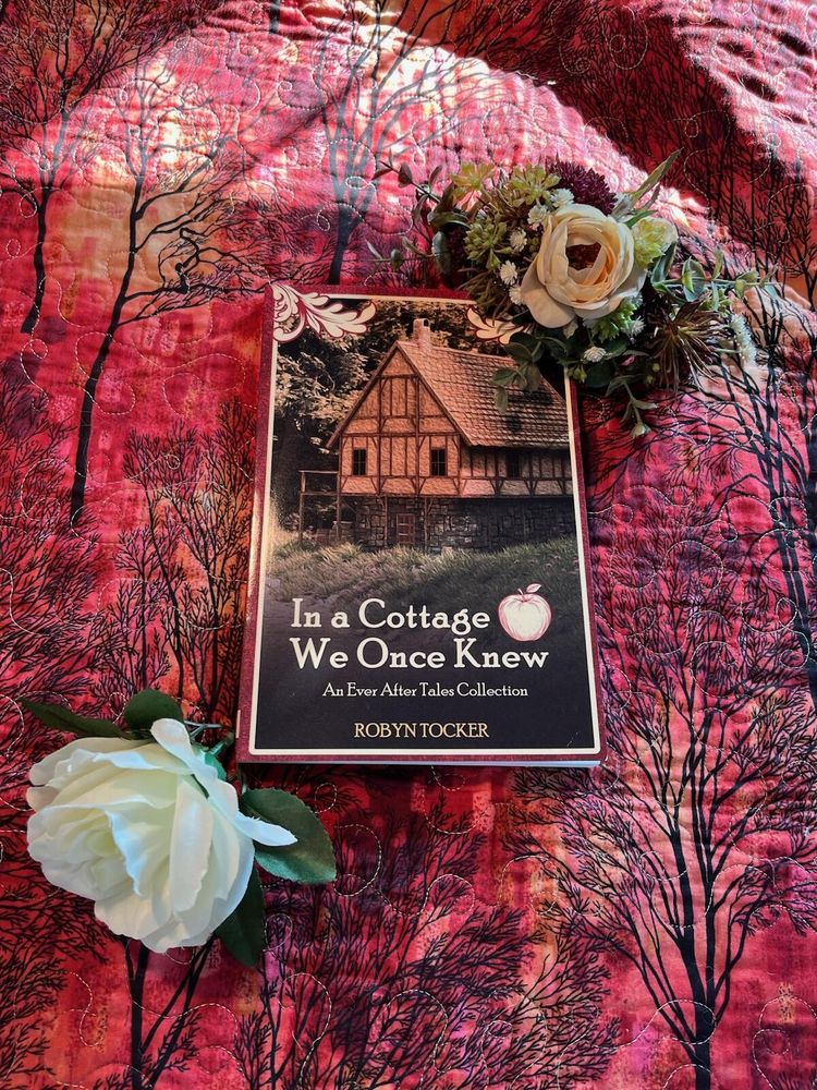 Beautiful cover of Robyn Tocker's In A Cottage We Once Knew. Deep red cover with picturesque wooden cottage and apple motif. Pictured on a red quilt with white roses.