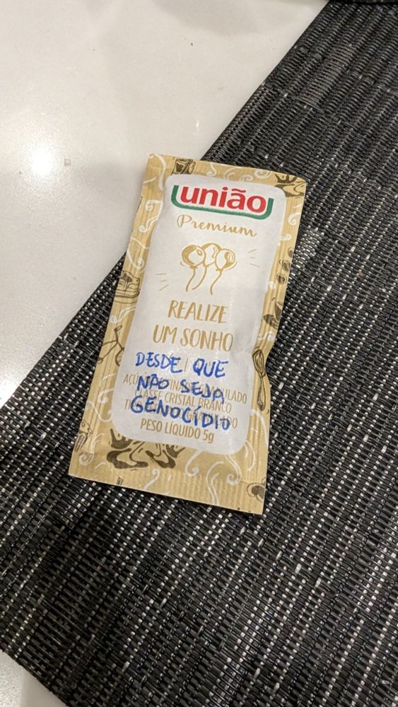 Embalagem de açúcar: "Realize um sonho". Anotado com caneta abaixo: "Desde que não seja genocídio".