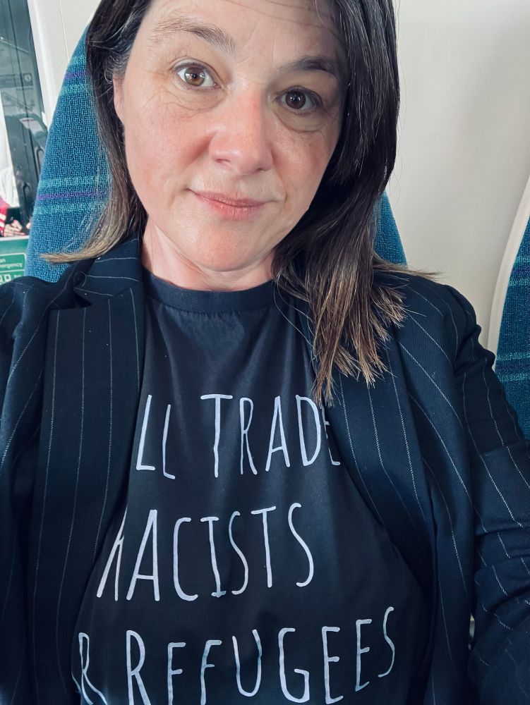 Lady #TypicalHistorian on a train on her way to teach on the Taliban’s destruction of Afghanistan’s music 1996-today, wearing a navy t-shirt that reads “will trade racists for refugees”