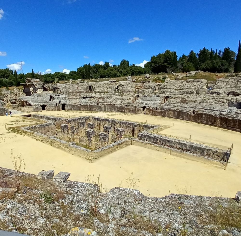 This picture shows the ruins of an old Roman amphitheater, which includes a sunken area in the center shaped like a cross. The sunken area has a series of pillars. There are trees and a deep blue sky in the background.