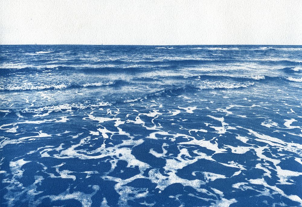 Photographie cyanotype de l'océan. Le ciel blanc sans nuage occupe le tiers supérieur de la photo, les deux tiers restants représentent l'ocean avec quelques vagues douces et de l'écume.