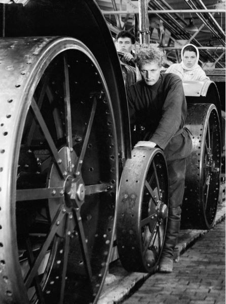 Photo en noir et blanc de trois personnes, a priori deux hommes et une femme, dans une usine soviétique de tracteurs, judicieusement placé(e)s derrières les roues immenses.