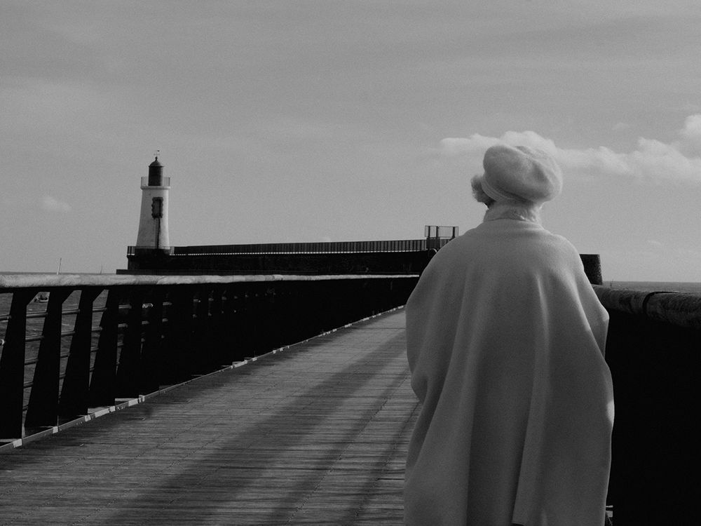 Photo en noir et blanc d'une dame âgée de dos, blanche et cheveux blancs, vêtue d'une cape en laine blanche et d'un béret blanc. Elle se trouve sur le quai qui emmène au phare.