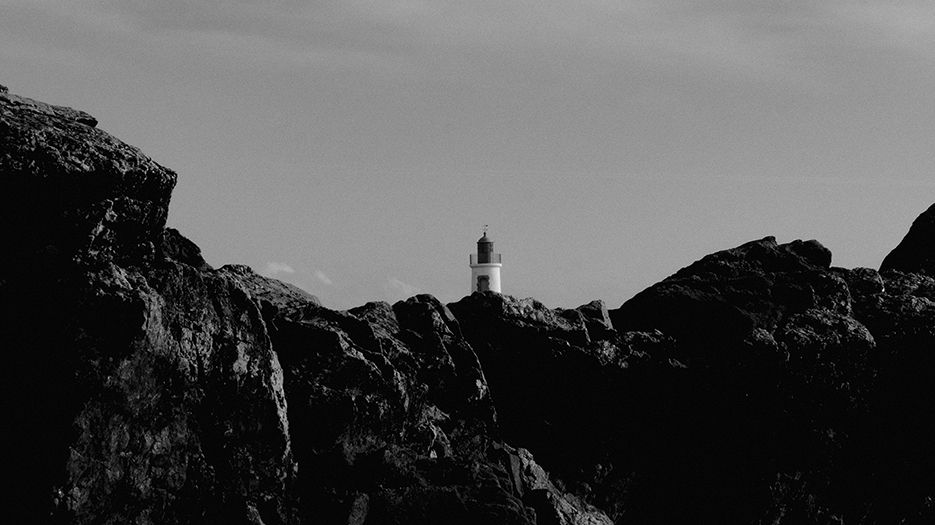 Photo en noir et blanc du phare de la Petite Jetée, aux Sables-d'Olonne, dont on n'aperçoit, au loin, que la tête surgissant de derrière un amas de rochers.