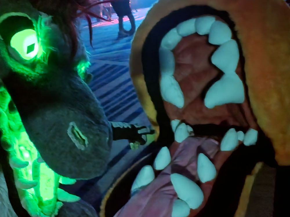 fursuit selfie of me crouched down by a giant maw, looking at the camera & pointing down its gullet