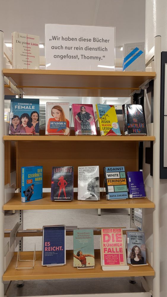 Auf einem Bücherregal steht ein Schild mit der Aufschrift "wir haben diese Bücher auch nur rein dienstlich angefasst, Tommy." Darunter eine Auswahl feministischer Bücher 