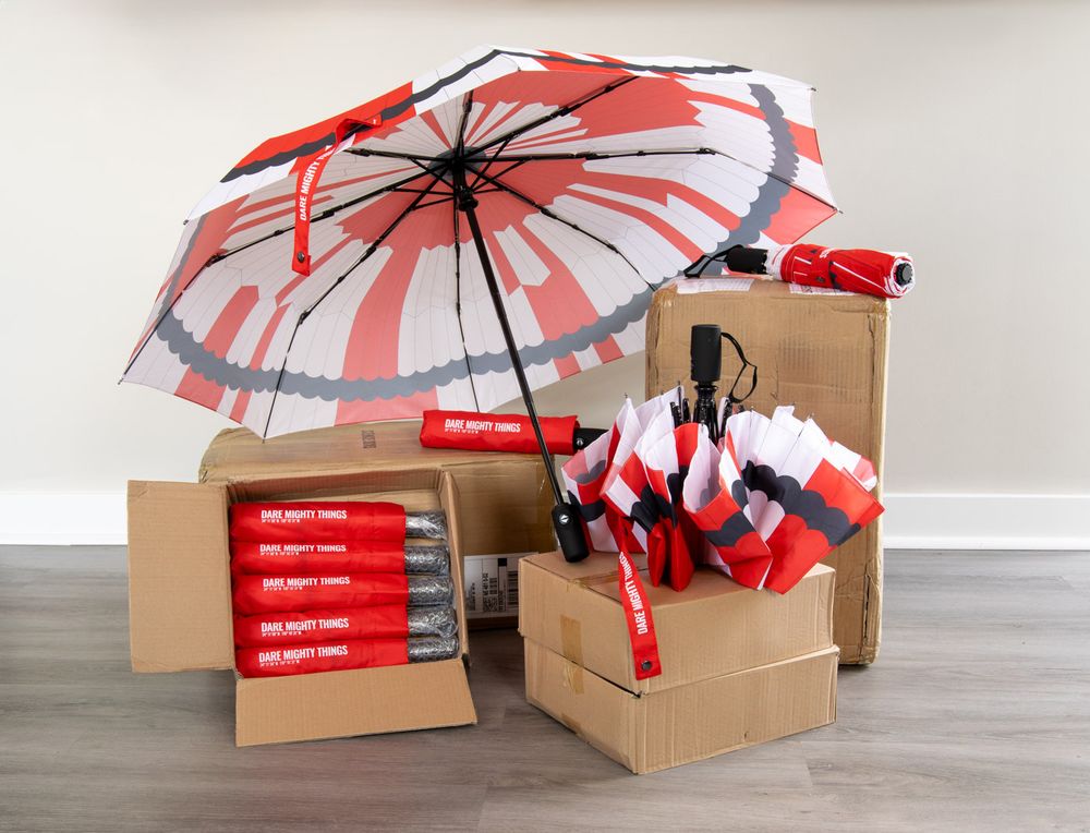 Boxes filled with Dare Mighty Things umbrellas. One is opened while others are closed and on display. Also several unopenend boxes can be seen.