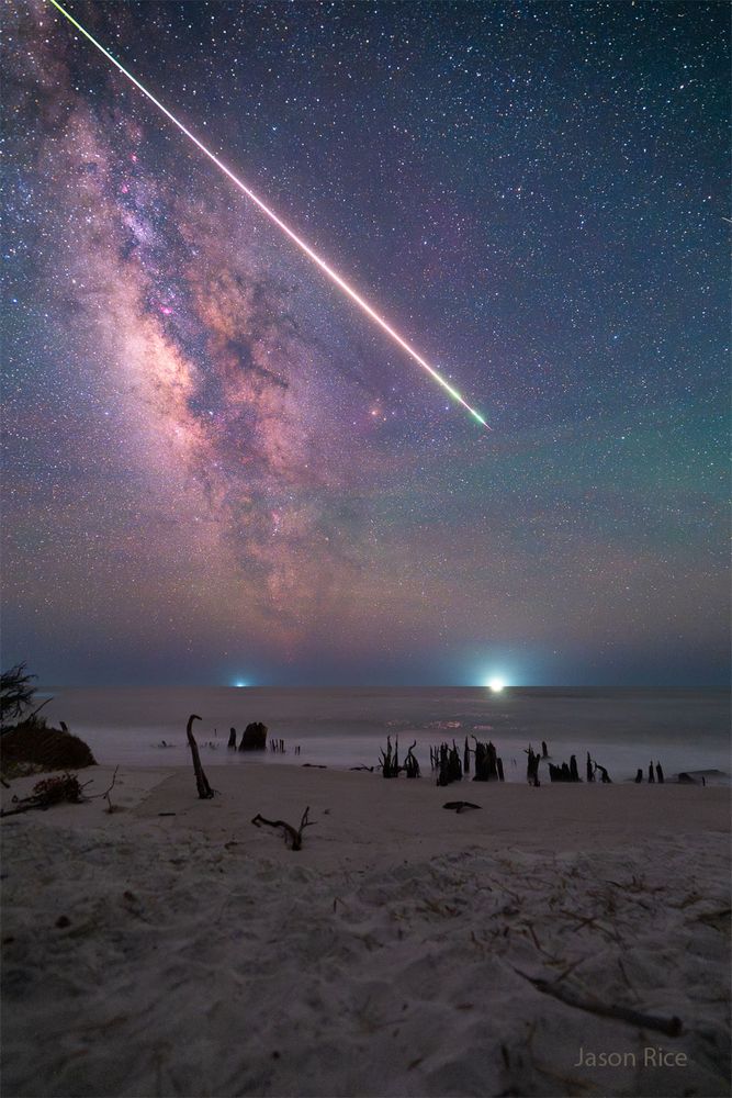 A bright streak is pictured through a starry sky over a 
beach spotted with the husks of dead trees. The rollover
shows the resulting smoke trail from the bright meteor.