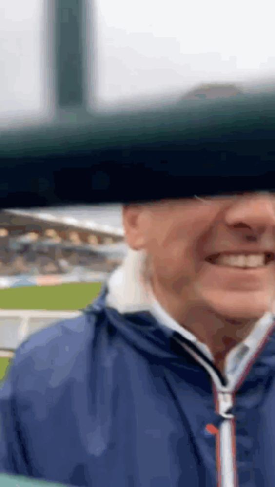 a man in a blue jacket is smiling in front of a baseball field