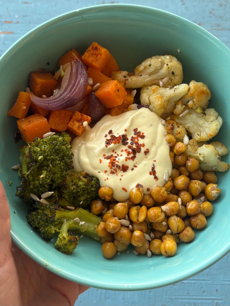 In einer türkisfarbenen Schüssel ist geröstetes Gemüse: Kichererbsen, Blumenkohl, Brokkoli, rote Zwiebeln und Süßkartoffeln. Darauf sind Sonnenblumenkerne gestreut. In der Mitte ist ein Klecks heller Dip mit Chiliflocken bestreut. 