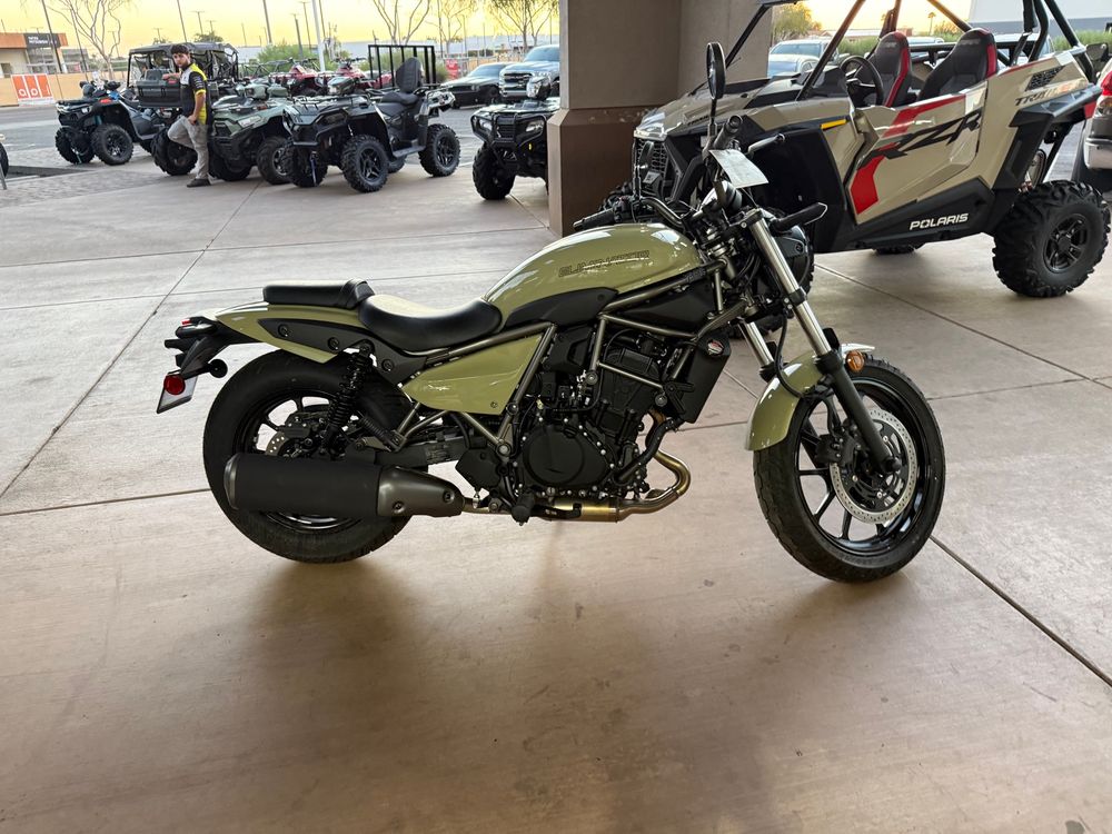 A photo of a pearl sand khaki colored Kawasaki Eliminator 450. It is parked at a power sports dealership, and you can see side-by-sides in the background 