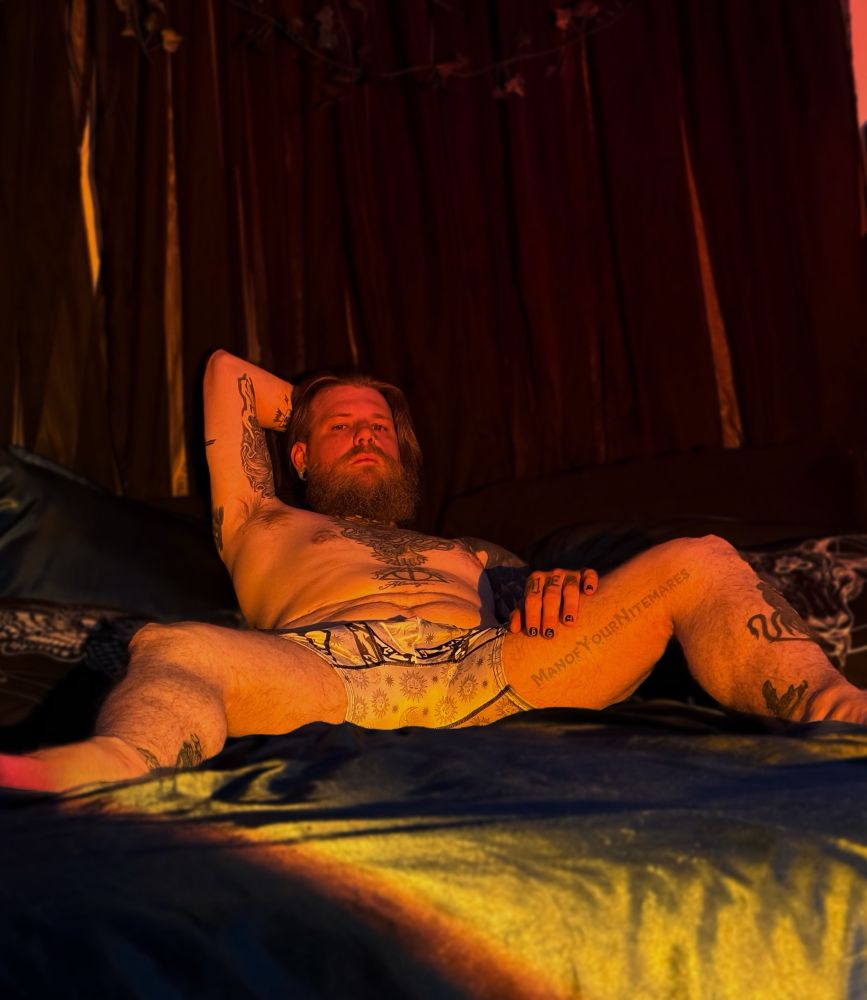 Nite is laid back with his legs spread open. He is wearing nothing except for gray boxers. He is staring intently at the camera, with one hand resting on his thigh, and the other behind his head