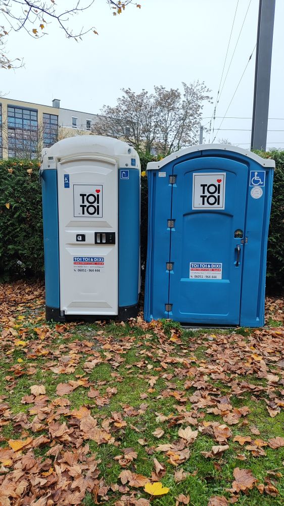 zwei mobiltoiletten aus blauem und weißem Kunststoff stehen auf einer Wiese mit braunem Laub