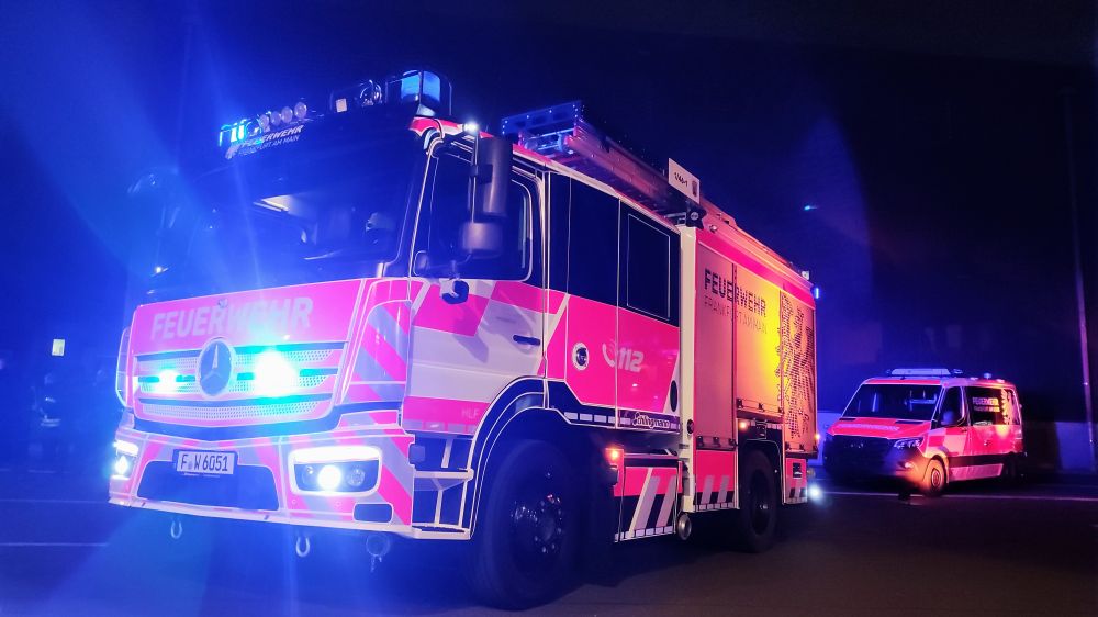 Ein Feuerwehrfahrzeug mit eingeschalteten blauen Warnlichtern steht nachts auf einer Straße. Das Fahrzeug ist rot-weiß lackiert und trägt die Aufschrift 'FEUERWEHR' sowie die Notrufnummer '112'. Dahinter ist ein weiteres Einsatzfahrzeug mit ebenfalls eingeschalteten blauen Warnlichtern zu sehen. Die Szene ist von blauem Lichtschein umgeben, was auf einen Einsatz hindeutet.

