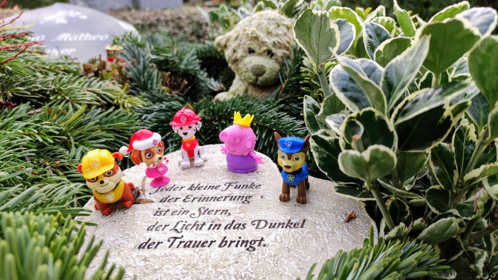 heller stein mit Sinnspruch, darauf kleine Plastikfiguren wie pepper pig und paw patrol. im hintergrund ein schwarzer grabstein für mateo und ein struppiger teddybär