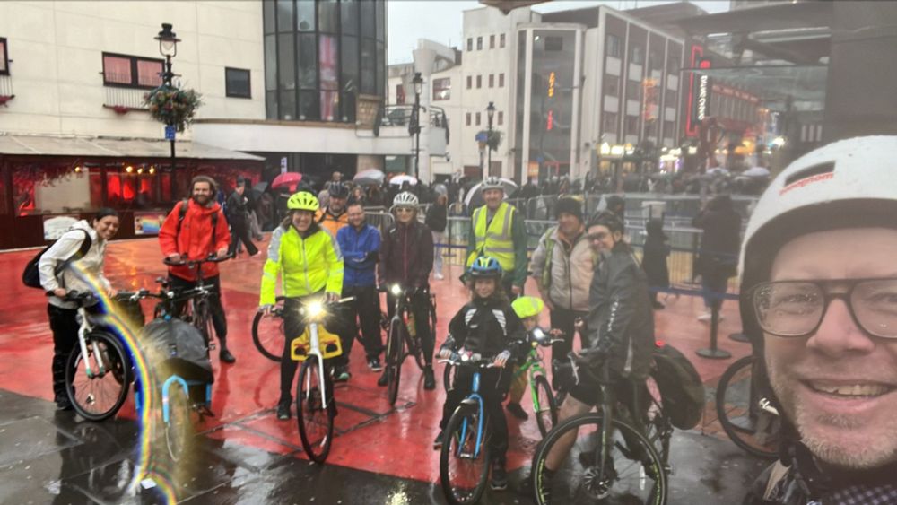A group of cyclists smiling in the rain during an evening ride through the city. Riders are wearing helmets, bright jackets, and waterproofs, with bikes of all sizes including children’s. Wet streets and umbrellas in the background show the rainy weather.