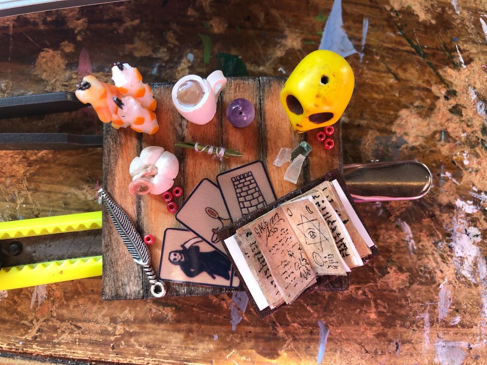 the hair clip from above. It's a wood work surface with candles, a cup of tea, a crystal ball, a yellow skull, red seed beads representing pomegranate seeds, some tiny bits of glass for crystals, an open grimoire, three tarot cards, a sage (rosemary) smudge stick, a white mini pumpkin, and a metal feather. 