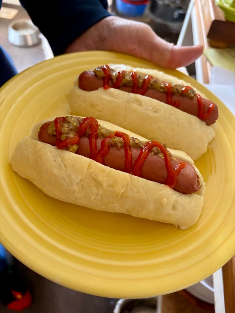 A hand holds a yellow Fiesta-ware plate containing two soft hot dog buns filled with Nathan’s 