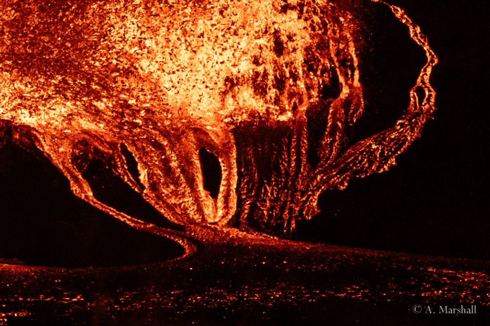 close up image of lava flowing away from the base of a fire fountain, feeding into a pond of lava. The night time view makes an extreme contrast between the red lava and the black rocks.