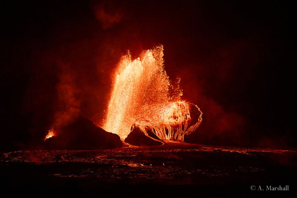 Large fire fountain shoots lava hundreds of feet in the air above a dark cone of rock. The night time view makes an extreme contrast between the red lava and the black rocks.