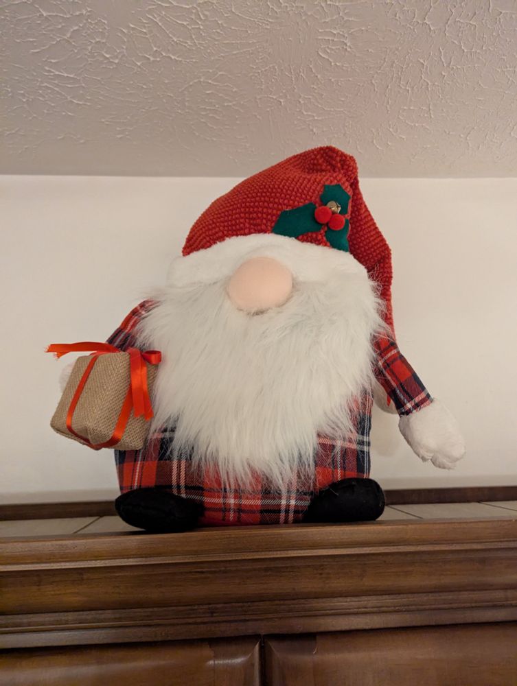 A Christmas gnome in red flannel with a red hat, that comes right down to his white beard, with a present in hand.