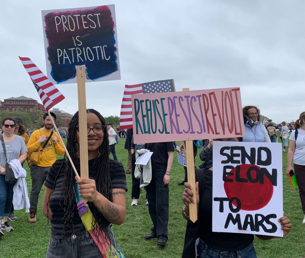 Two women hold signs and a flag next to the Washington Monument, with people visible in the background. One sign reads “Protest is patriotic.” Another says, Refuseresistrevolt.” And a third has a large red dot with “Send Elon to Mars” superimposed over it.