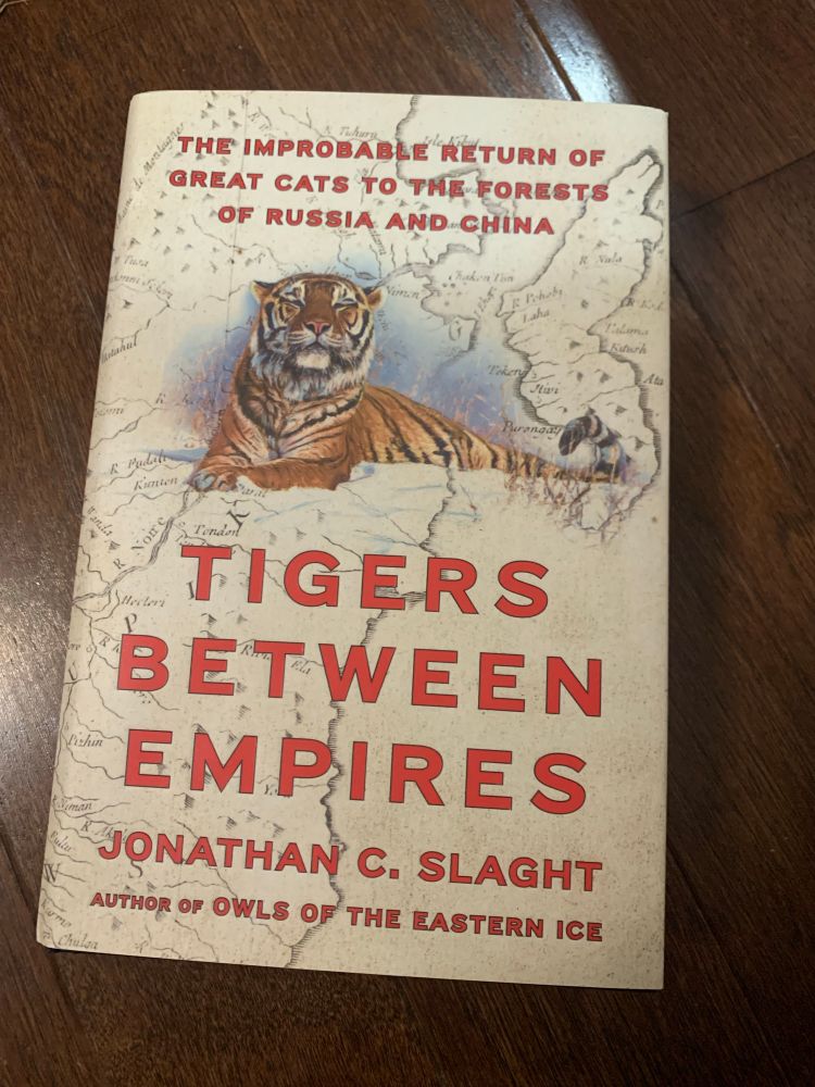A photo of a hardcover book titled TIGERS BETWEEN EMPIRES by Jonathan C. Slaght. The subtitle is “The improbable return of great cats to the forests of Russia and China.” The background is a map of Eastern Asia with a bright orange-and-black tiger lying in the snow