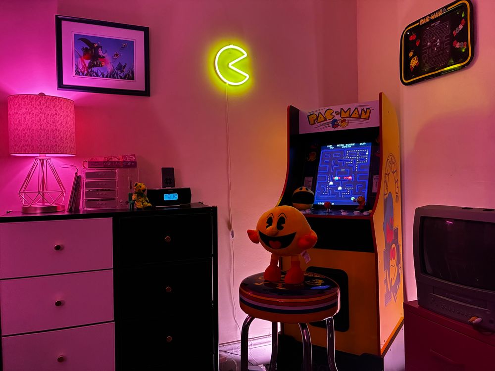 Corner of room with Pac-Man plush on Pac-Man stool with Pac-Man machine behind. To the left, a black and white dresser with an iPod in ihome atop and some books. Above it, a lithograph of a bugs life. To the right, a CRT tv atop a pink file cabinet. 