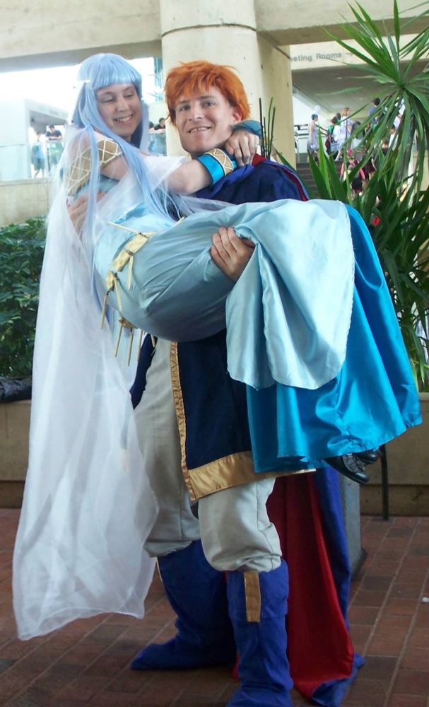 Two people cosplaying Eliwood and Ninian from the videogame Fire Emblem: Blazing Sword. Eliwood is holding Ninian in a bridal carry; they are both smiling.