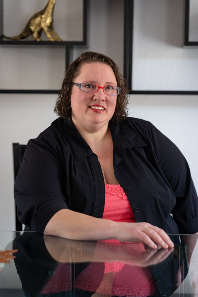 A fat, white woman with chin length curly hair and pink and grey glasses smiles in an open black button down with a pink lace trim cami underneath. She is seated at a glass table with one arm resting on the table. There is a black geometric shelf behind her with a gold figurine, her reflection can be seen on the table.