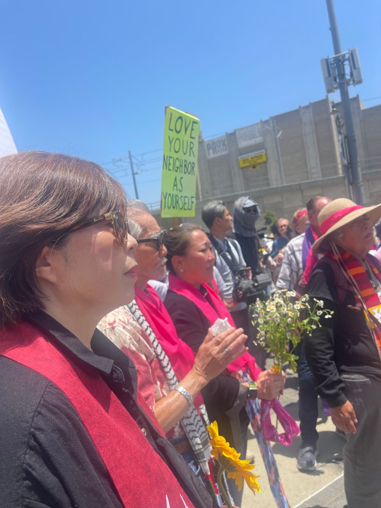 A group of people stand in a peaceful outdoor protest under a clear blue sky. A person in the foreground holds a sunflower, and another has daisies. A bright green sign reads, "Love your neighbor as yourself."