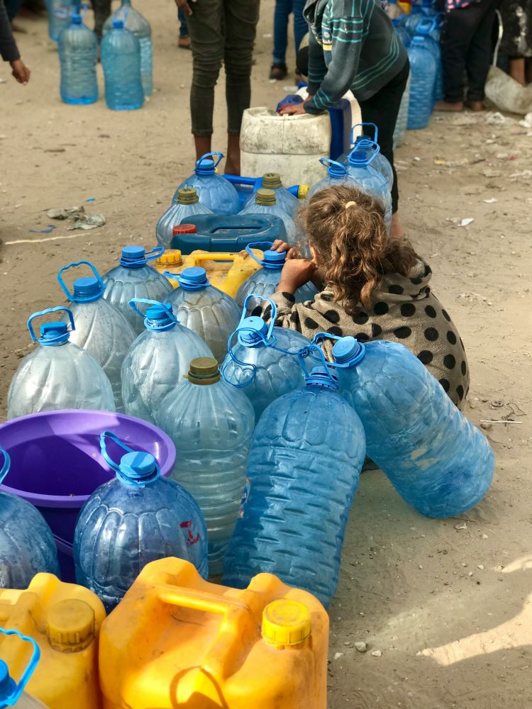 A young girl in Gaza sits among empty water jugs, symbolizing the ongoing struggle to find food and potable water in the region. Photo by Emad El Byed's via Unsplash. 