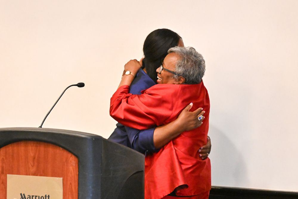 The Rev. CeCe Armstrong and Dr. Phyllis W. Sanders embrace behind a podium as Armstrong accepts her award. 