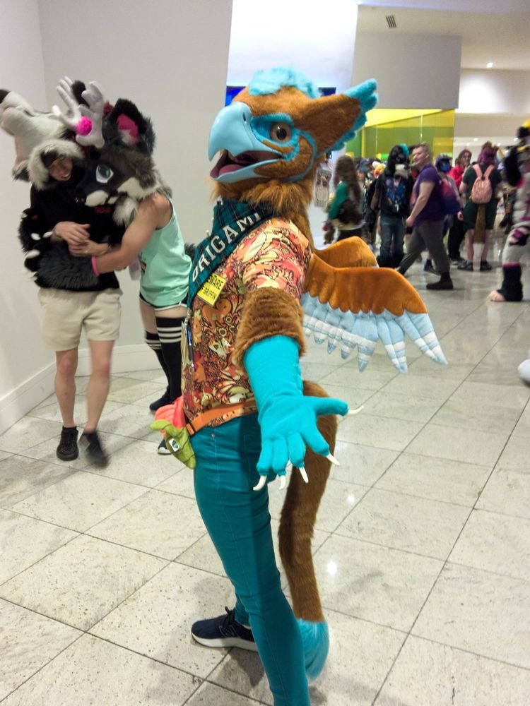 A full body shot of me in my gryphon partial. I'm standing sideways, beak wide open and left hand held out, palm up. There are other people and fursuiters in the background.