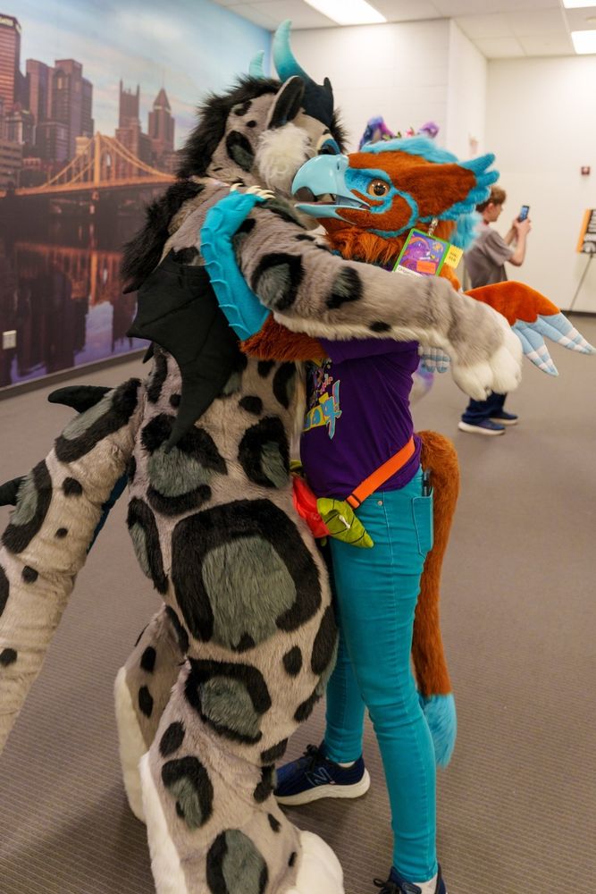 Me in my gryphon partial fursuit hugging a snow leopard dragon fursuiter. She is taller than me and drapes her arms over my shoulders. My arms are wrapped around her shoulders from underneath her arms.