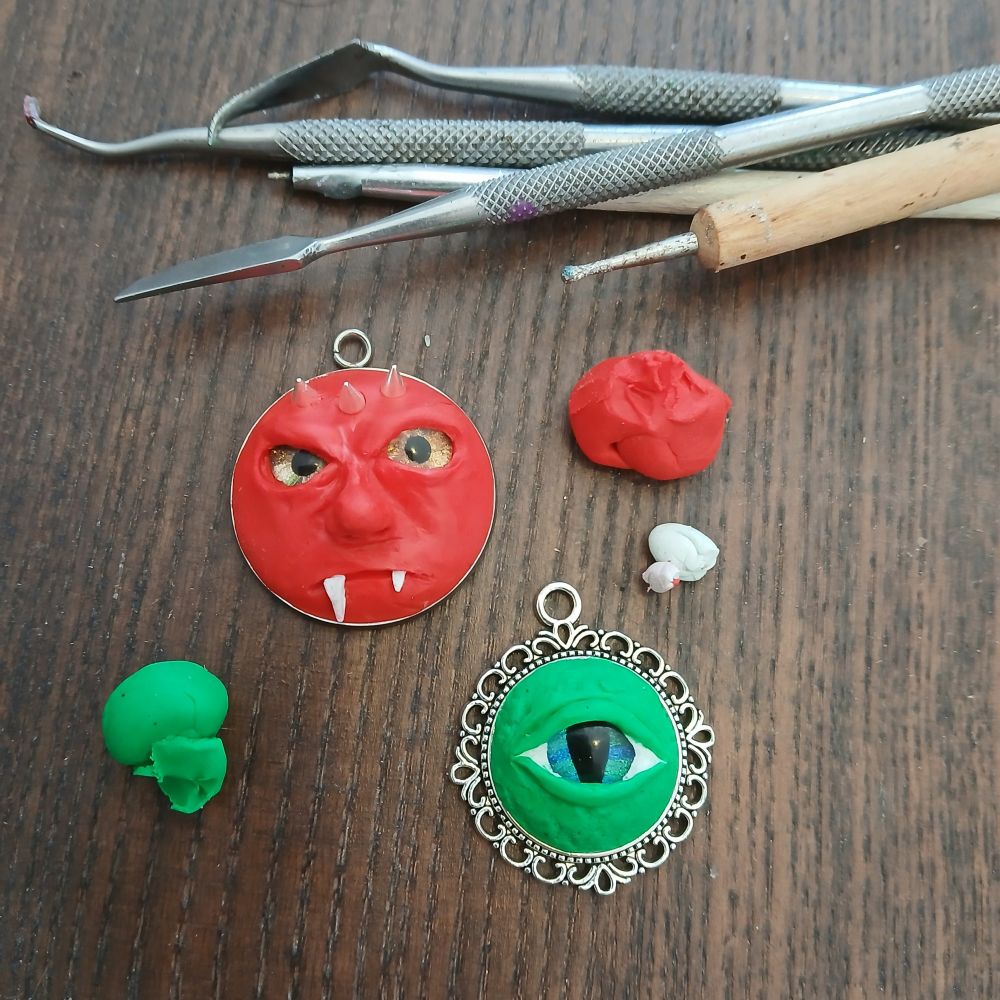 Two unfinished polymer clay pendants, one a red frowning face with three spikes in the forehead and prominent fangs, and the other a small staring eye in green clay surf texturing around the eye. There are also stray bits of clay and well-used sculpting tools in shot. 