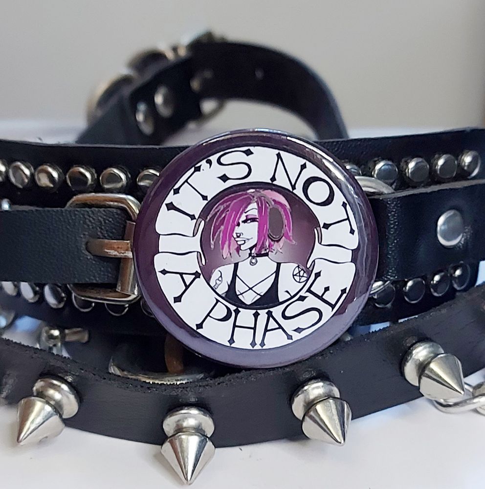A badge sitting in front of some leather accessories that says "It's not a phase" and has a cute alt girl in headphones in the middle. 