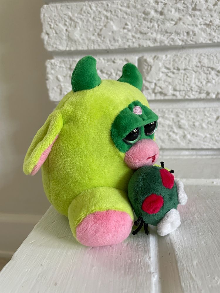green plushie with tongue sticking out that is meant to evoke a furby and a frog. a small fly plushie is attached to it.