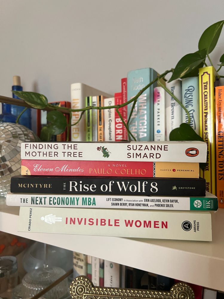 A stack of five books on a white shelf, partially surrounded by trailing green leaves. Titles include Finding the Mother Tree by Suzanne Simard, Eleven Minutes by Paulo Coelho, The Rise of Wolf 8 by Rick McIntyre, The Next Economy MBA, and Invisible Women by Caroline Criado Perez. A colorful bookshelf is blurred in the background.