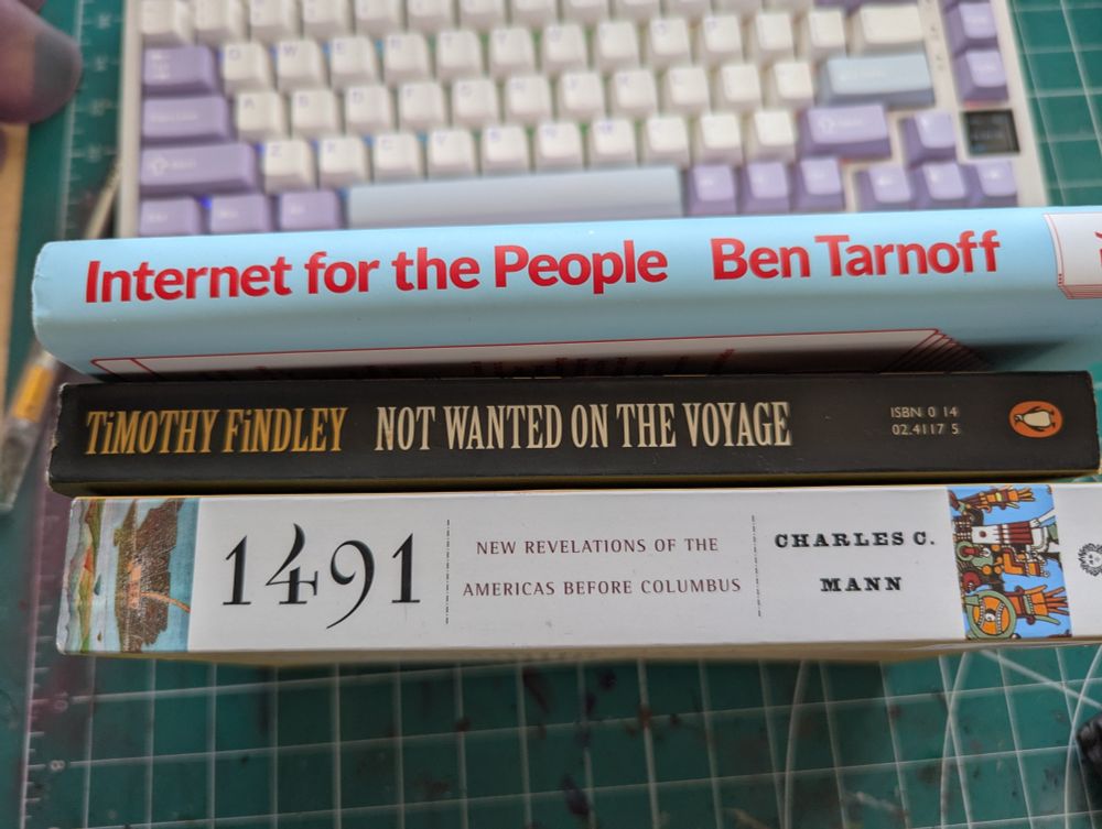 Three books stacked on a green cutting mat in front of a pastel mechanical keyboard. Titles include Internet for the People by Ben Tarnoff, Not Wanted on the Voyage by Timothy Findley, and 1491: New Revelations of the Americas Before Columbus by Charles C. Mann.