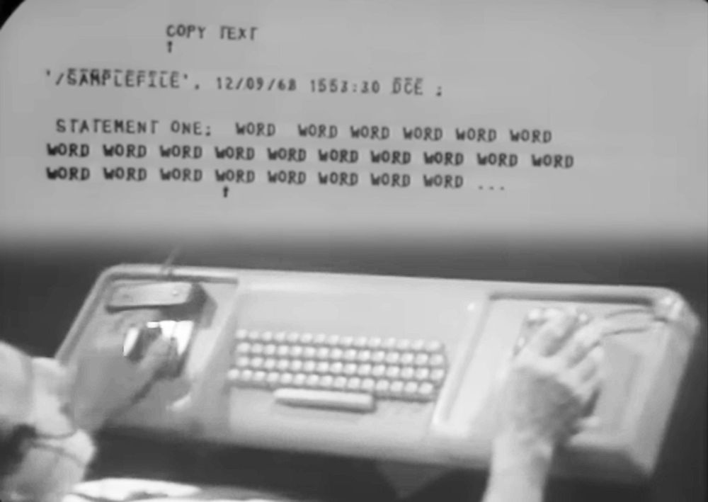 An image from Engelbart's demo. At the top, the screen shows lines of text. The bottom of the image shows Engelbart at a special desk. The desk has a keyboard in the center with a keyset on the left and a mouse on the right.