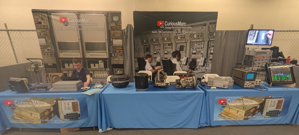 Photo of a booth at OpenSauce. Three tables are covered with vintage electronics of various sorts. Mike is working on the Apollo CDU at the left.
