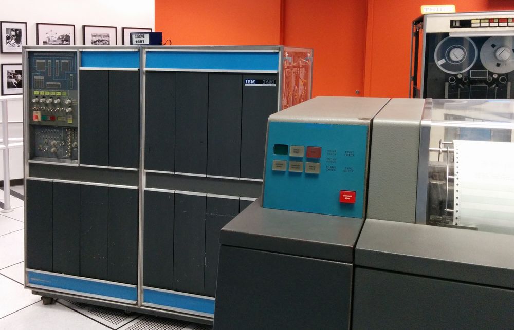 The IBM 1401 is a vintage computer about the size of two refrigerators. It has dark gray panels with blue and chrome trim. In the upper left, a control panel has numerous lights and buttons. The large line printer is visible in the foreground. A tape drive with two reels of tape is in the background.