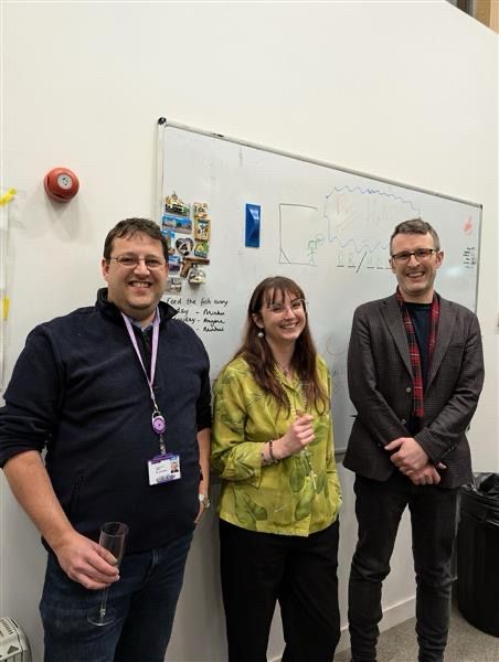 A joyful PhD student holding a glass of champagne is posing with her thesis examiners after successful defence of her work.