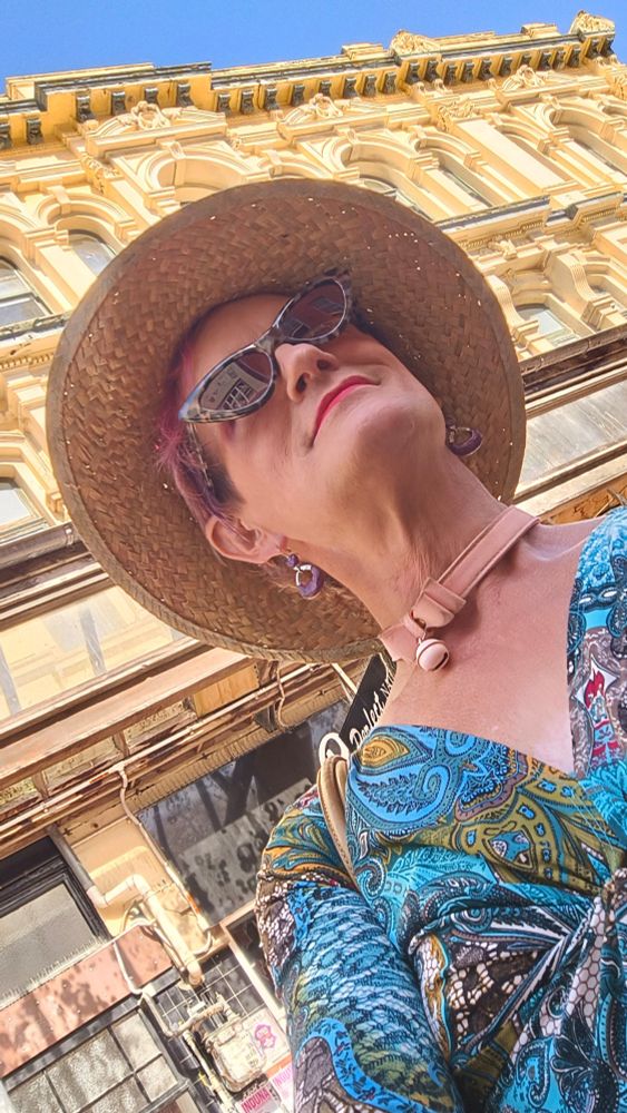 Margie Fancypants, wearing a tropical straw hat and paisley blue top and cat-eye sunglasses, before an elaborate Rococo façade in the late afternoon summer sunlight on Cuba Street, Wellington, New Zealand.