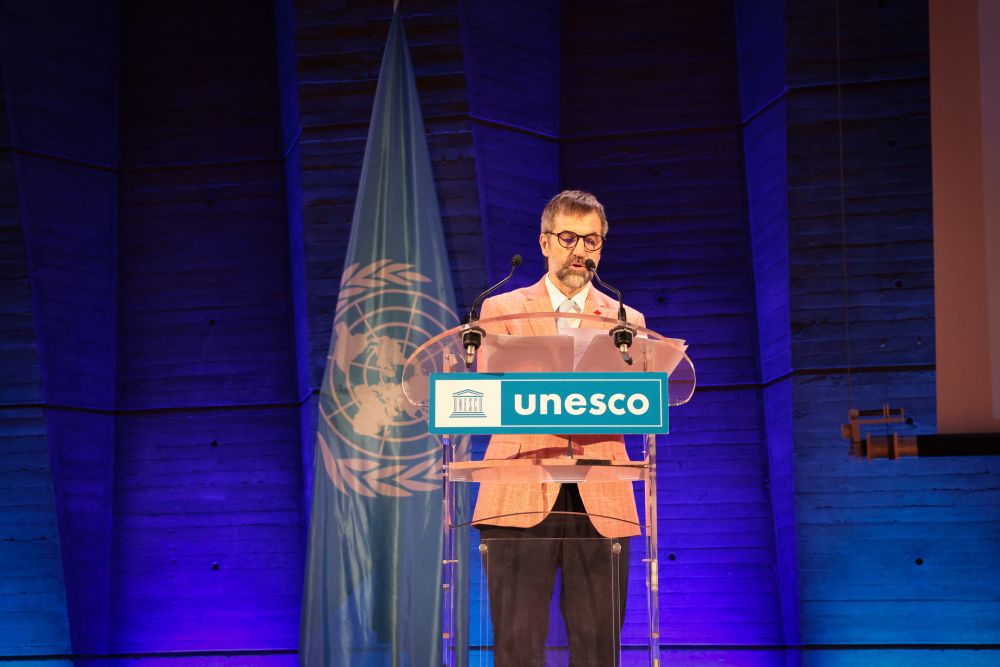 Le ministre Guilbeault se tient derrière un podium et prononce un discours. Le drapeau de l’UNESCO est à l’arrière plan.