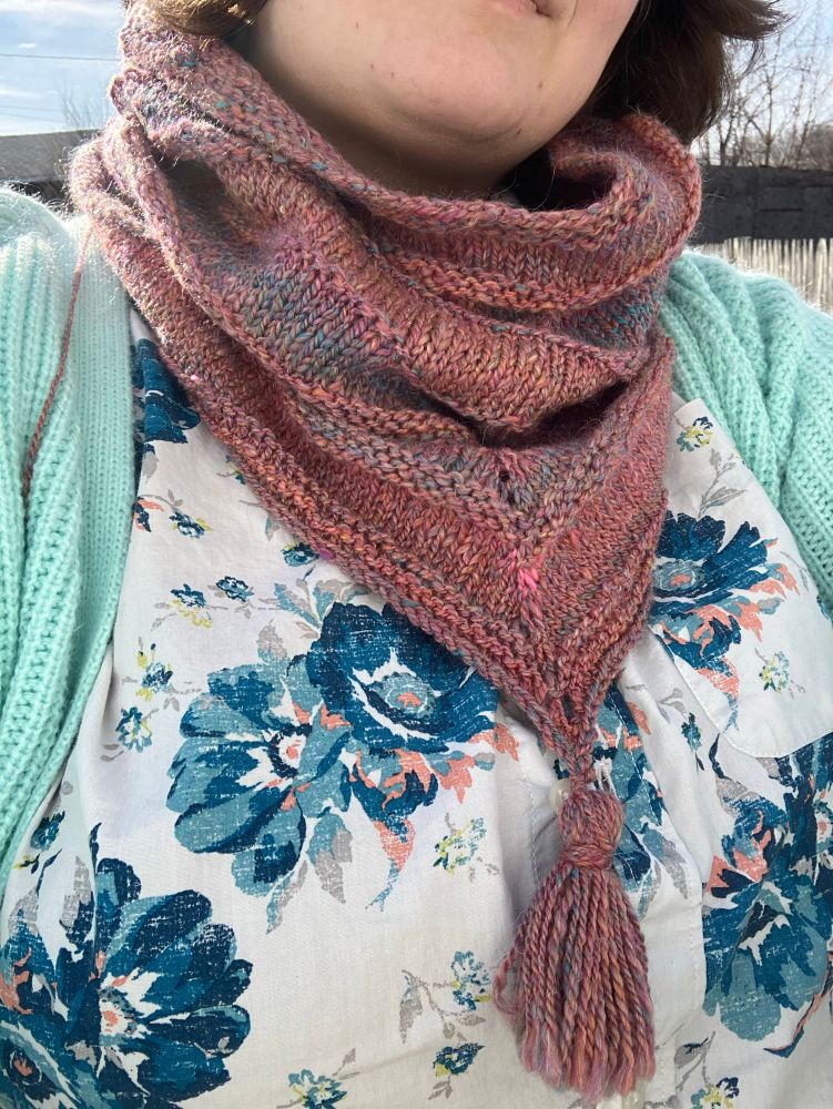 A person in a white shirt with blue flowers wearing a dusty pink cowl knit from handspun yarn. Subtle bands of blue and orange appear naturally in the yarn. The cowl is slouchy, with occasional purl stripes throughout. It comes to a sharp point in front, with a tassel of the same yarn attached to the tip. 