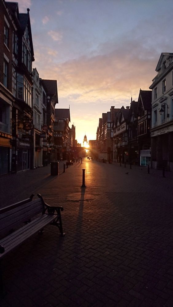 Very early sunrise over Chester's Foregate, quite far away, with a bench and a bollard in the foreground, throwing shadows 